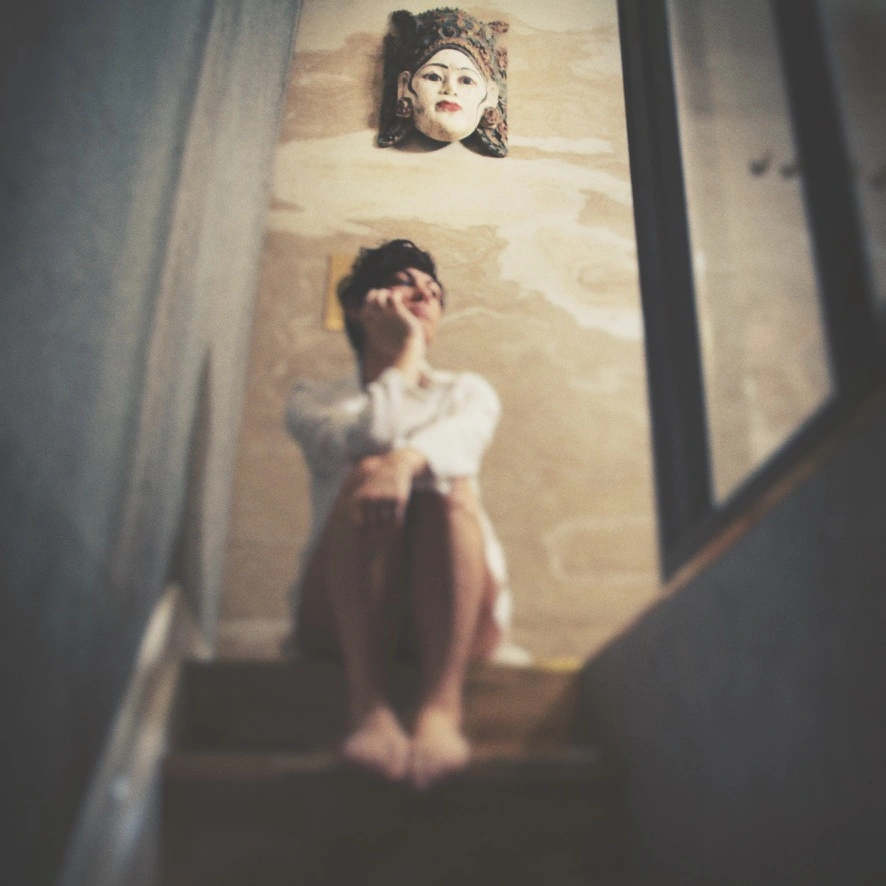 Une silhouette assise dans l’escalier, sous un masque balinais, dans une photographie au flou artistique.